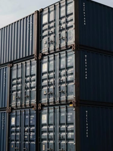 Close-up graphic composition of steel shipping containers stacked in a deep-water port under crisp overcast daylight, high-contrast metallic textures.