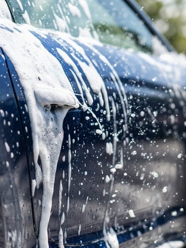 Crisp commercial photography of thick active foam sliding down a clean dark blue sports car panel under bright daylight, capturing high-speed water droplets, 50mm