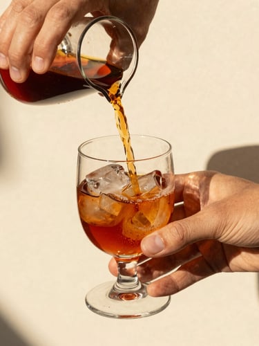 Close-up of hands pouring deep terracotta-colored liqueur into an elegant glass with ice, bright high-contrast Mediterranean sunlight, sharp shadows, warm editorial photography, 35mm