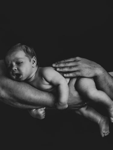 Black and white newborn photography of a father's hands cradling a sleeping baby against a dark background.