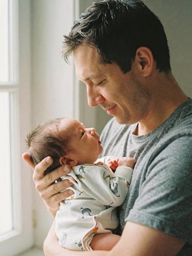 A tired but smiling father holding his newborn baby close to his chest under soft morning window light, warm domestic atmosphere, 35mm film look