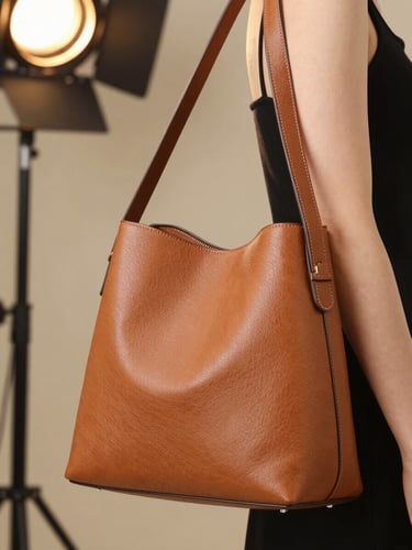 Close portrait-oriented editorial shot of a structured caramel leather shoulder bag hanging from a woman's shoulder, warm studio light from above-left, soft bokeh background in warm ivory tones, visible texture of the leather strap, no face visible