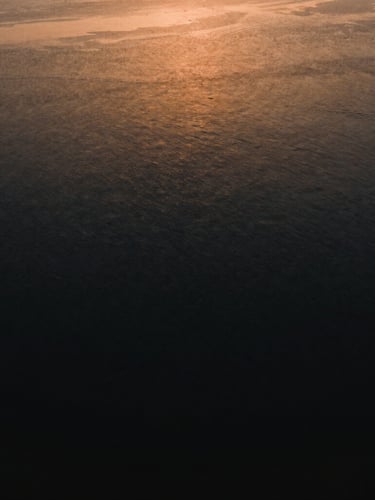 Cinematic wide angle drone view of Marano Lagunare lagoon, quiet dark water, golden hour light reflecting on the surface, deep shadows, rich ochre tones, minimalist composition, 35mm anamorphic lens
