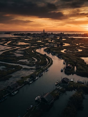 Wide cinematic drone shot of Marano Lagunare lagoon at golden hour, deep moody shadows, rich ochre and charcoal tones, 35mm anamorphic lens