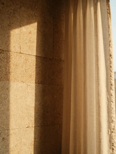 Close-up of ancient hand-laid limestone walls meeting smooth modern linen drapery, warm golden-hour light filtering through a tall window, architectural detail.