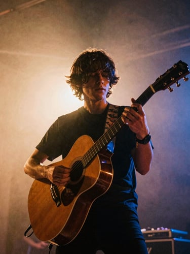 Low-angle dynamic shot of a musician playing an acoustic guitar on stage, warm amber backlighting, stage haze, 35mm film grain