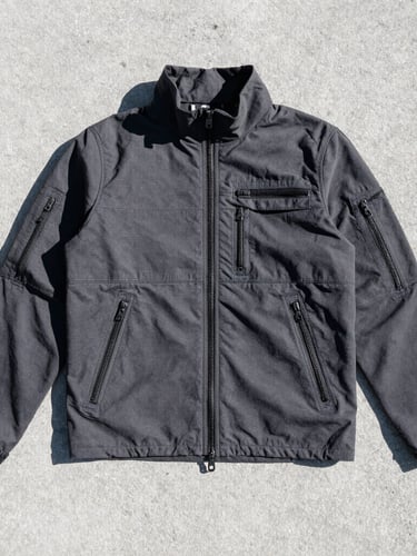 Stark technical streetwear flat-lay on cold concrete, high-contrast direct sunlight, sharp focus on heavy zippers and reinforced stitching, desaturated tones