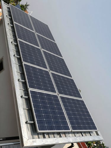 Wide environmental shot from ground level looking up at a fully installed residential rooftop solar array on a flat-roofed Bihar home, galvanized mounting structure visible, overcast daylight revealing panel texture and wiring quality, no people, expansive sky to the right