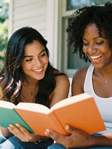 Candid medium-close up of a smiling Latina woman and Black woman sitting together on a sunny porch, holding colorful books, laughing, bright warm natural light, 35mm film look.