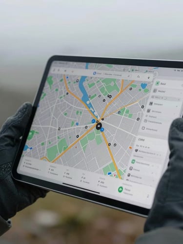 A close-up photograph of a gloved hand holding a modern tablet displaying a map with precise GPS coordinate markers, overcast outdoor lighting, cool tones