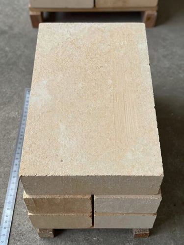 Close overhead flat-lay of stacked high-alumina refractory fire bricks on a concrete warehouse floor, controlled fluorescent warehouse light, sharp texture detail showing the dense grain and cream-buff surface color, ruler partially visible at edge for scale reference