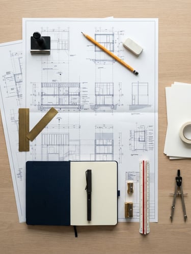 Overhead flat-lay of high-quality stationery, a yellow graphite pencil, and structured architectural blueprints on a clean white desk, bright north-facing daylight, sharp focus, 35mm