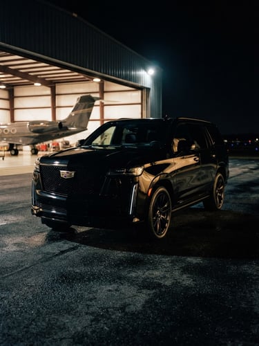 Low-angle heroic shot of a polished all-black 2026 Cadillac Escalade parked outside a private aviation hangar at night, gold reflections, shallow depth of field, 35mm