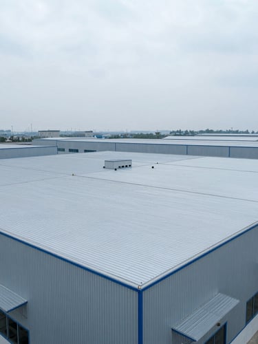 Wide-angle aerial perspective of a massive modern industrial warehouse roof with clean metal seams, crisp overcast daylight, cool metallic blue tones, 35mm