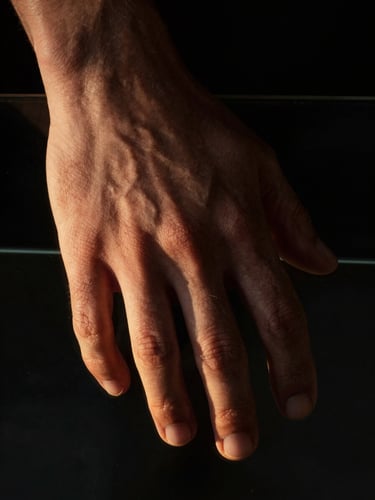 Extreme close-up of a human hand resting on dark glass, dramatic side-lighting from the left casting half the hand in deep shadow, the fingertips barely catching a thin streak of warm golden light, macro framing, psychological weight, black background