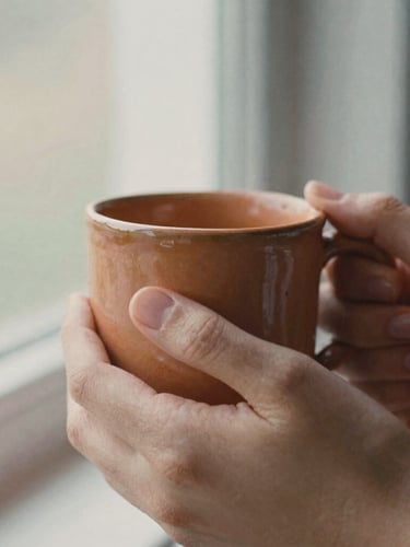 Close-up detail of hands holding a warm terracotta ceramic mug, soft morning light filtering through a window, shallow depth of field, warm editorial 35mm film style