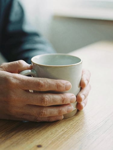 A close-up of hands wrapping a warm ceramic mug on a wooden table, soft diffused overcast daylight, muted tones, shallow depth of field