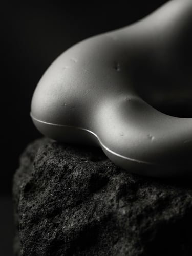 Extreme close-up of a smooth, matte platinum-silicone sculptural form resting on a raw dark stone plinth, dramatic low-key chiaroscuro lighting, deep shadows.