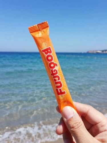 Close-up action shot of a hand holding a vibrant orange Fuego Rollypop tube against a bright blue Mediterranean sea background, direct midday coastal sunlight, sharp focus