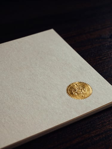 Tight macro shot of a heavy, textured cream-colored paper document with a subtle gold foil embossed seal, placed on a dark walnut desk, moody side lighting with deep shadows.