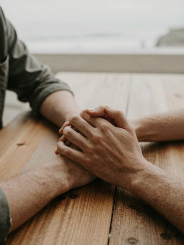 A candid, warm close-up of two people holding hands over a wooden table, soft Pacific Northwest overcast daylight, muted earthy tones, 35mm.