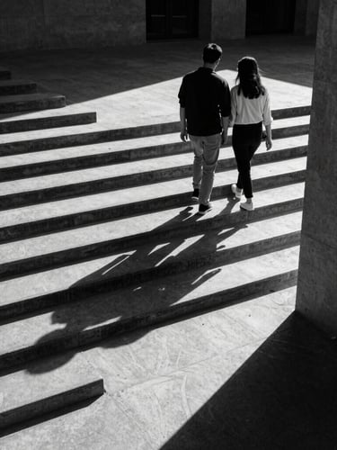 A high-contrast medium-format black and white photograph of a couple walking down a concrete staircase in harsh sunlight, long shadows casting geometric patterns, architectural framing, candid movement.