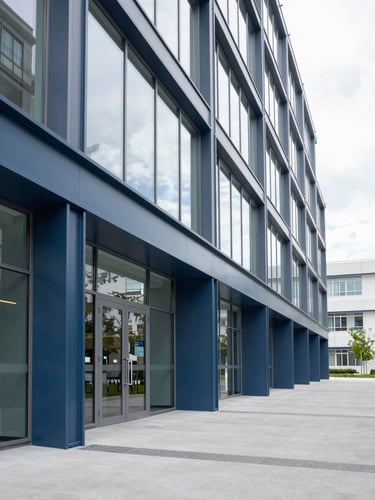 A wide, clean, daylight-filled architectural detail of a modern medical center corridor, showcasing geometric glass panels, structural steel, and deep slate blue accents with generous negative space.