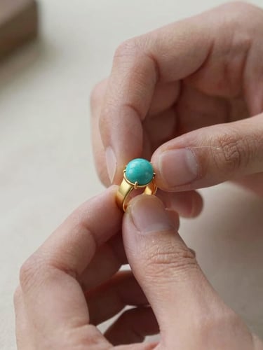 An extreme close-up of a jeweler's hands carefully setting a raw turquoise stone into a matte gold band, soft natural light, shallow depth of field