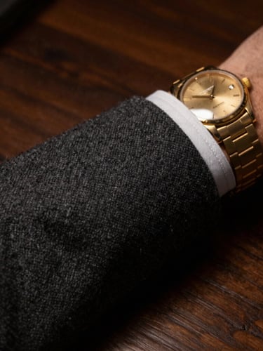 Tight macro detail of a tailored charcoal wool cuff and a gold watch against a dark wood table, low-key warm lighting, atmospheric