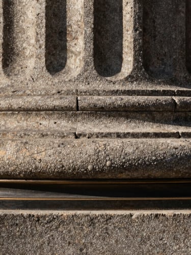 Macro close-up of heavy textured architectural stone column meeting a dark bronze metal frame, dramatic side-lighting, deep shadows.