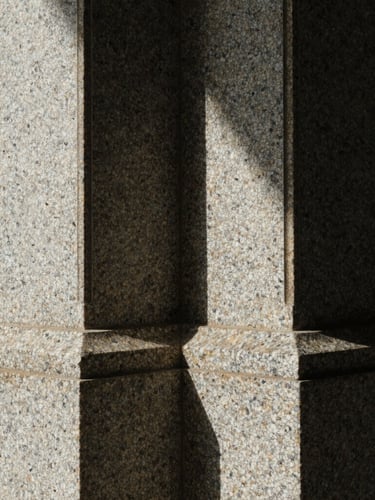 Macro close-up of a heavy, textured granite structural column, dramatic side-lighting casting deep shadows, architectural security.