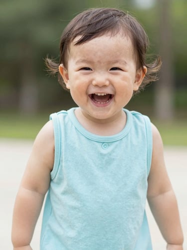 Bright candid portrait of a happy toddler laughing in a soft pastel blue cotton romper, natural outdoor daylight, 35mm