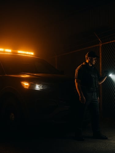 Low-angle, high-contrast night shot of a marked patrol vehicle with active amber lightbars illuminating a dark industrial fence, a tactical officer in clean utility uniform inspecting the perimeter with a flashlight.