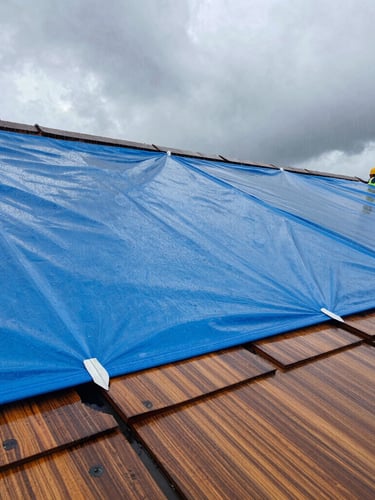 Close-up of industrial waterproof blue tarping being secured tightly over damaged roof shingles during heavy rain, overcast dramatic sky, professional safety gear visible