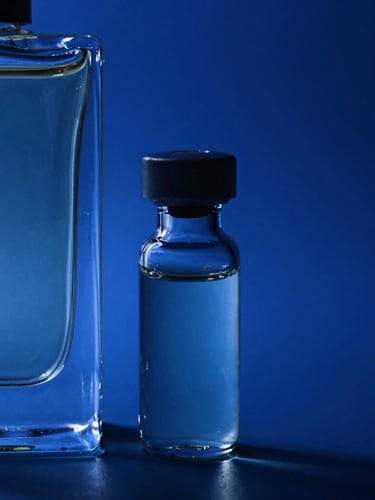Extreme macro shot of a sleek glass decant vial next to a luxury perfume bottle, sharp liquid refraction under dramatic royal blue studio lighting, deep shadows.
