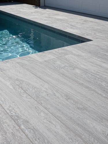 A low-angle shot of a luxury backyard pool deck finished in a modern slate-gray textured coating, bright afternoon sunlight reflecting off the water, clean lines, high-contrast cool tones.