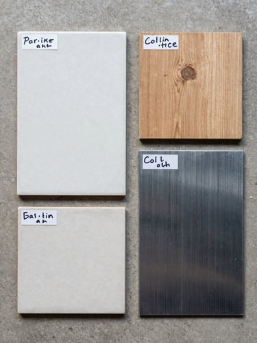 Overhead flat-lay of material samples arranged on a concrete surface — ceramic tile sections, a timber plank cross-section, cold-rolled steel sheet — with handwritten spec labels visible, even diffused daylight from above, no shadows, authentic material textures clearly visible