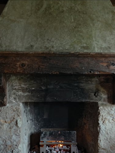 Close-up of original 18th-century dark timber beams and a deep stone hearth with a soft glowing ember, moody editorial photography, 35mm.
