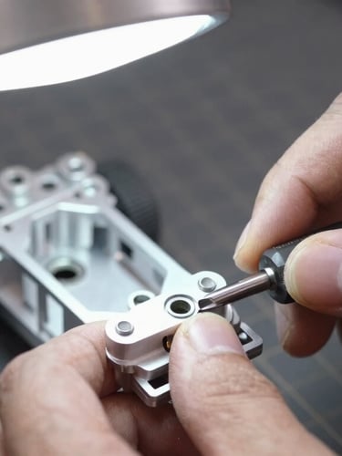 Close-up macro shot of a mechanic's hands using a hex driver on an RC aluminum chassis, bright overhead workshop light, dark grey cutting mat background, sharp macro details, 50mm lens