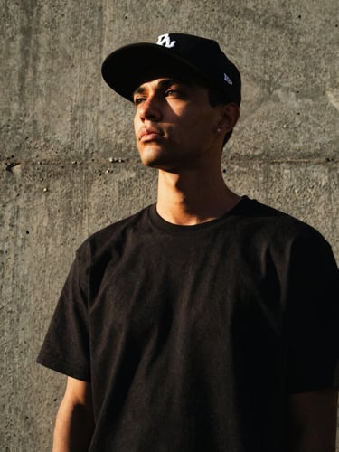 Low-angle portrait of a streetwear model wearing a structured black snapback cap against a raw concrete wall in Karachi, dramatic high-contrast lighting, 35mm film style