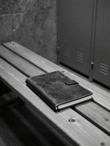 Editorial black and white photography, dramatic natural light, deep shadows, high contrast, 35mm film grain. A closed leather-bound notebook resting on a wooden bench inside an empty, dark concrete locker room.