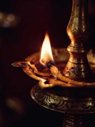 Macro close-up of a traditional Kerala brass oil lamp glowing with warm flickering light, deep maroon shadows, antique gold reflections, 35mm photography