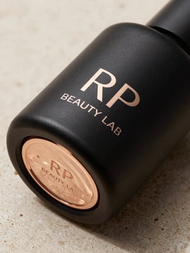 Macro studio shot of a sculptural matte black RP Beauty Lab bottle on a raw beige stone surface, directional light reflecting champagne gold accents, high-end cosmetics photography