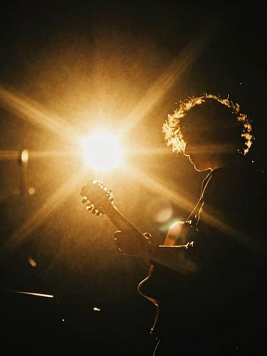 A dramatic backlit portrait of a musician holding a guitar, warm golden-hour light flares cutting through deep black negative space, high-contrast luminous editorial photography, 35mm