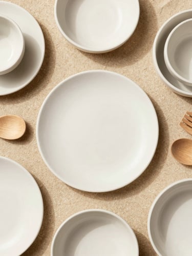 Overhead flat-lay of curated ceramic tableware and minimalist wooden kitchen utensils on a warm sand surface, bright diffused natural daylight, soft shadows, portrait aspect.