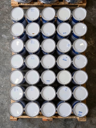 Close overhead flat-lay on a warehouse pallet, stacks of sealed paint drums and shrink-wrapped tool crates photographed from directly above under even warehouse fill-light, origin labels visible on carton faces, concrete floor visible at the edges