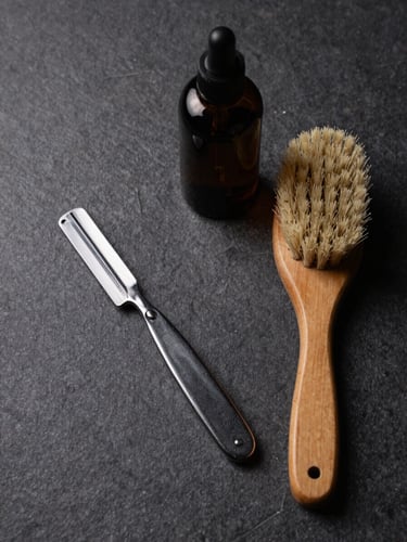 Overhead flatlay of grooming tools — a straight razor, a wood-handled brush, and a dark glass bottle on a matte stone surface, studio strobe lighting, high contrast, minimal negative space