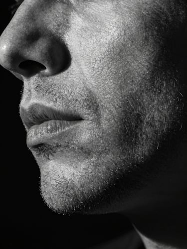 Extreme close-up of a man's jawline and beard texture, dramatic rim lighting from the left casting deep shadow, fine beard hair detail visible against dark studio background, high-contrast monochrome editorial feel