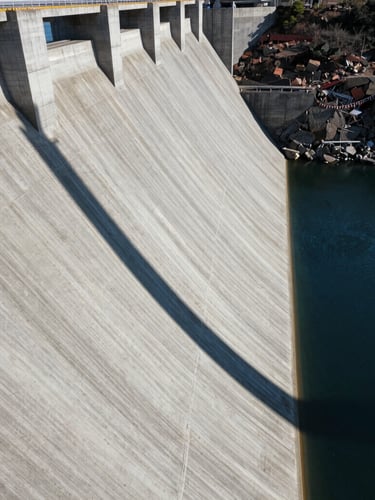 Vista aérea de una presa de hormigón masiva bajo la luz solar intensa del mediodía, sombras geométricas marcadas, agua azul profundo a un lado, textura rugosa del cemento.