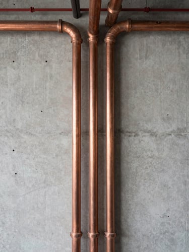 A straight-on technical photograph of clean, organized copper and PVC pipes running along a concrete wall in a modern commercial utility bay, bright natural lighting, architectural layout feel.